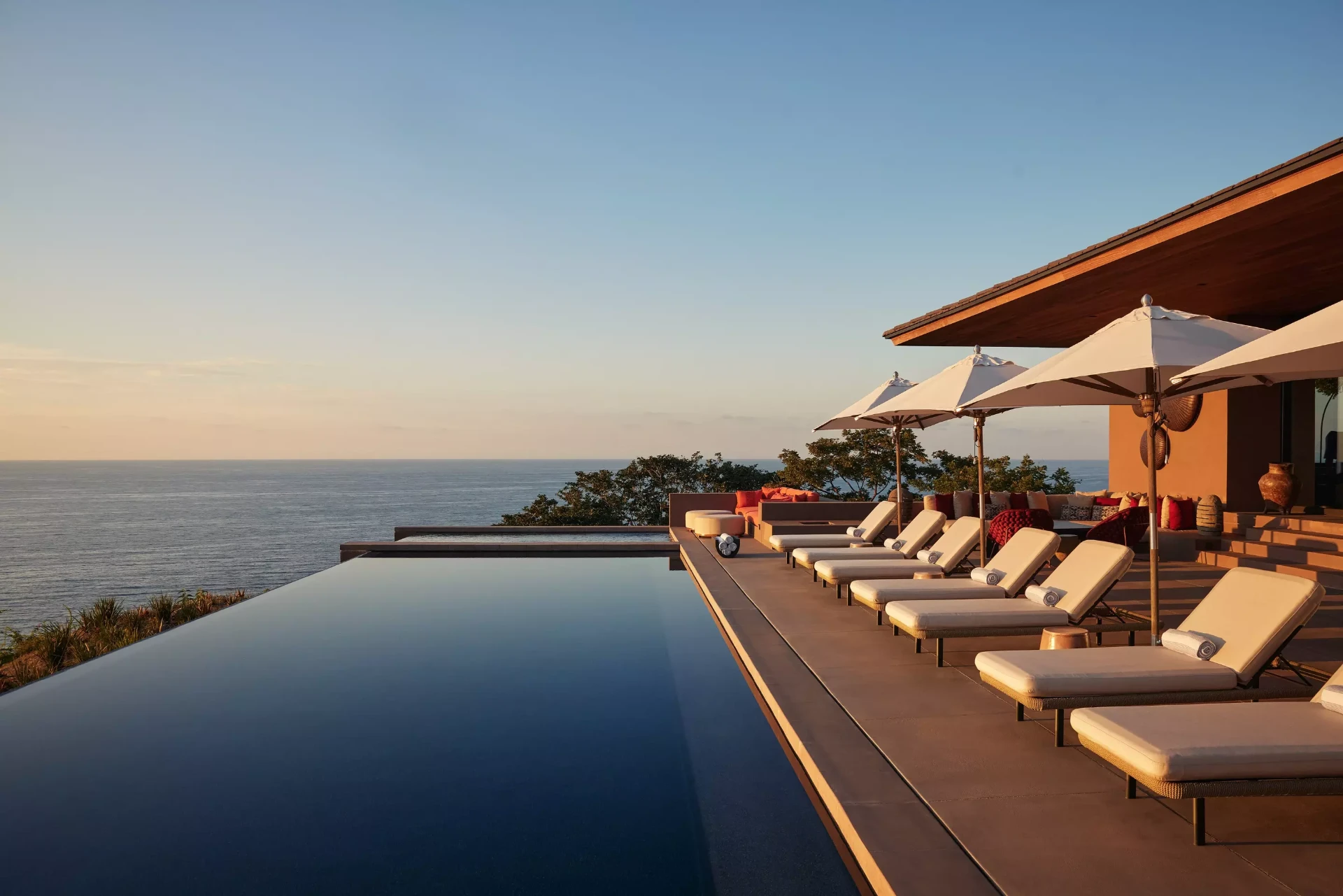 Sun loungers and parasols on a terrace beside an infinity pool, with the sea beyond