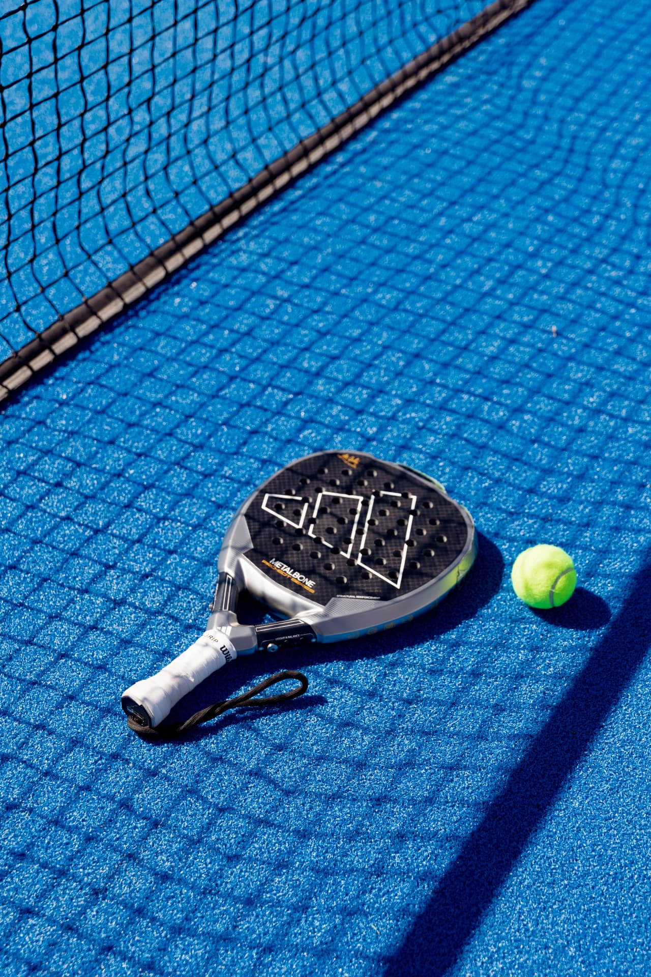 A padel racket and a neon yellow ball resting on a blue padel court in the shadow of the net