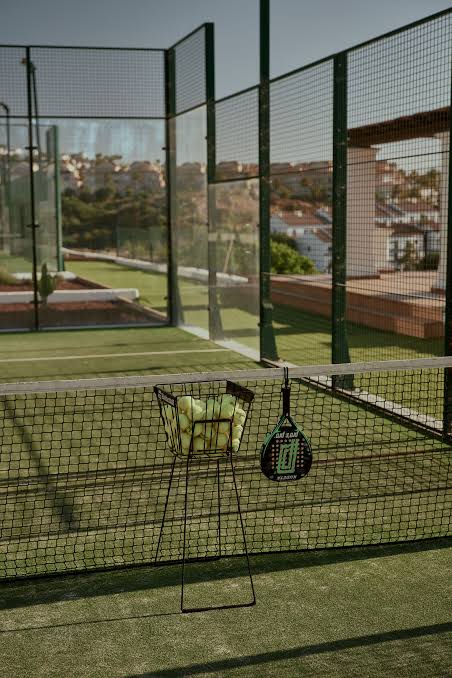 A ball basket and a racket on a green padel court at OKU Andalusia, with hillside houses beyond the fencing